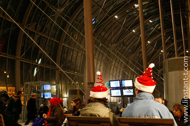 Deux personnes de dos, avec un bonnet de Père Noël , assises sur un banc de la grande verrière de la gare de Strasbourg Bonnets-03.jpg